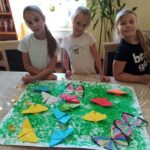 zajęcia w bibliotece dzieci przy stole prezentują swoją pracę duży arkusz pomalowany na zielono i motyle