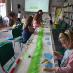 dzieci i bibliotekarka na długim stole wyklejają papierową łąkę