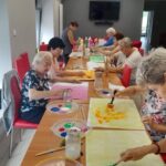 Seniorzy seidzą przy stole i malują