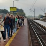 Grupa młodzieży stoi na peronie, w tle widać dworzec kolejowy