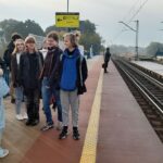 Grupa młodzieży stoi na peronie, w tle widać dworzec kolejowy