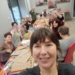 grupa seniorów siedzi przy stole i działa artystycznie, na pierwszym planie twarz prowadzącej zajęcia (selfie)