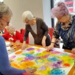 warsztaty artystyczne dla seniorów, uczestnicy pracują przy stole nad kolorową planszą
