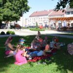 dzieci siedzą na kocach na trawie i słuchają bajki czytanej przez bibliotekarkę