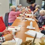 grupa Seniorów przy wspólnym stole maluje farbą deseczki
