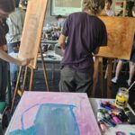 Warsztaty malarskie, młodzież maluje przy stołach i sztalugach, na pirewszym planie płotno w kolorach różowym i niebieskim