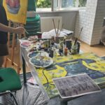 Warsztaty malarskie, młodzież maluje przy stołach i sztalugach, na pierwszym planie obrazy