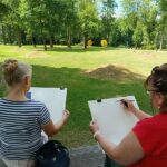 park, dwie kobiety siedzą na ławce tyłem do nas, malują, widzimy ich prace