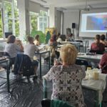 w sali siedzą osoby na krzesłach przy stolikach, warsztaty malarskie