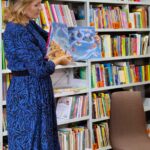 bibliotekarka pokazuje otwartą książkę, stoi w tle regał z książkami
