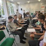 kilkanaście młodych ludzi siedzi w dwóch rzędach twarzami do siebie, malują się nawzajem