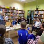 w bibliotece, bibliotekarka czyta dzieciom i dorosłym siedzącym wokół