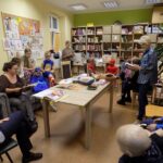 w bibliotece, uczestnicy spotkania czytają dzieciom i dorosłym siedzącym wokół