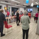 grupa osób stoi przy krzesłach i wykonuje ćwiczenia gimnastyczne