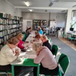 grupa seniorów siedzi przy stołach i rozwiązuje zadania