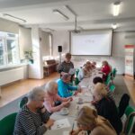 grupa seniorów siedzi przy stołach i rozwiązuje zadania