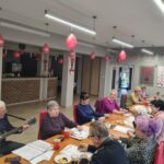 grupa osób siedzi przy stołach i rozwiązują zadania pisemne śpiewając piosenkę