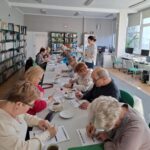 grupa seniorów siedzi przy stołach i rozwiązuje zadania