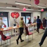 grupa osób wykonuje ćwiczenia na stojąco przy krzesłach.