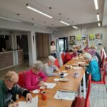 grupa seniorów siedzi przy stołach i rozwiązują zadania na pamięć i koncentrację