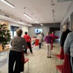 grupa seniorów wykonuje gimnastykę na stojąco przy krzesełkach