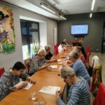 grupa seniorów siedzi przy stołach i rozwiazuja zadania