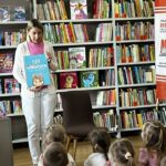 bibliotekarka pokazuje książkę, w tle regały z książkami